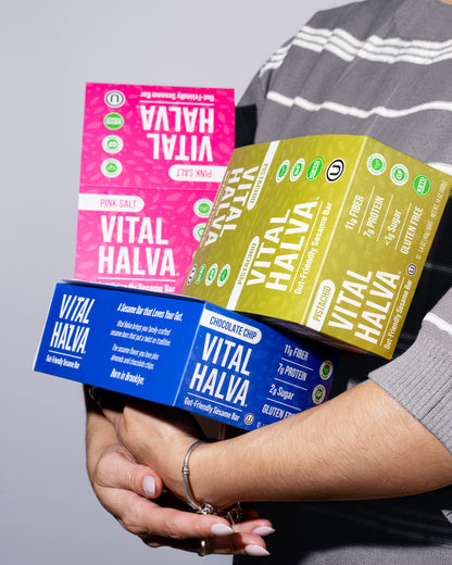 A person in a gray striped shirt holds three colorful Vital Halva sesame snack bar boxes stacked together against a plain light gray background. The boxes are pink, blue, and olive green with white text.